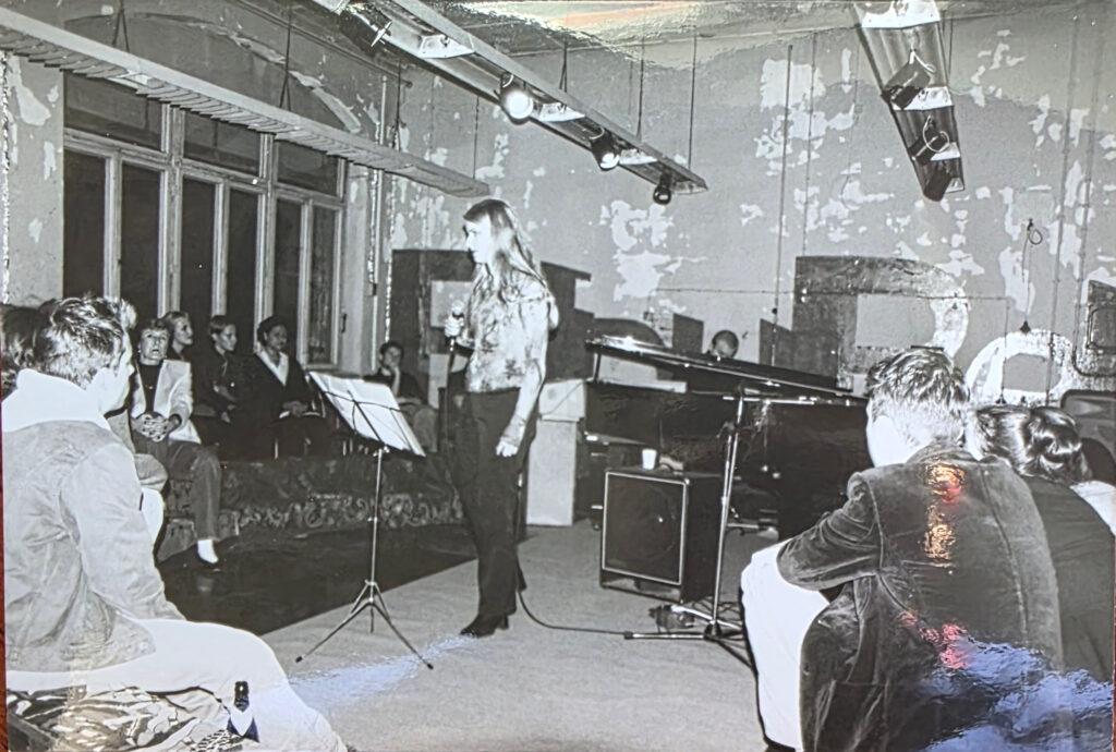 Kristina Tuomi performing at BLAUBETON Kulturetage in Berlin with a pianist, surrounded by the audience and the central performance space.