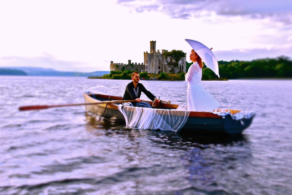 Kian Lee and an actress dressed in white with a white parasol in a rowing boat on the water during a concept-video shoot in Ireland, with a castle ruin in the background