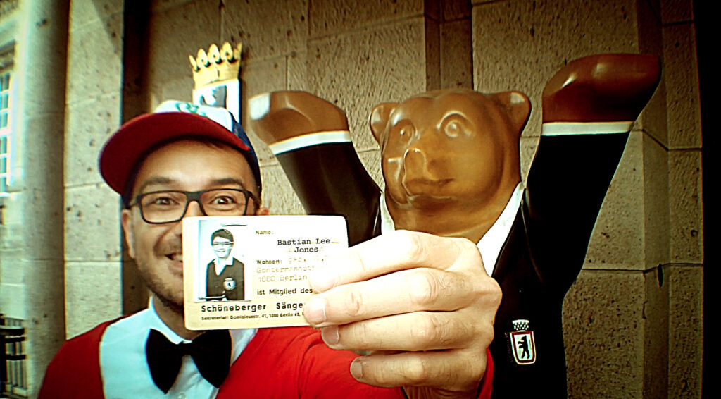 Bastian Lee Jones holding his historic Schöneberger Sängerknaben choir card in front of a Berlin Buddy Bear.